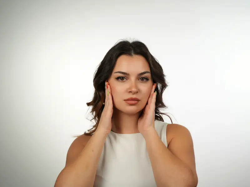 Woman touching her cheeks while looking at the camera, suggesting evaluation of facial laxity and jawline definition.