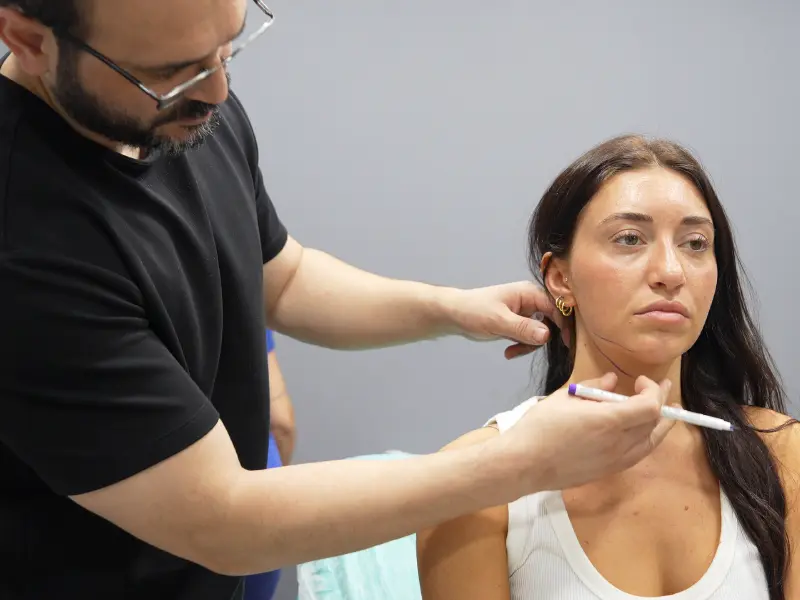 Dr. Akif Mehmetoğlu using a surgical marker to draw incision lines on a female patient's neck during a pre-op consultation at AKM Clinic, demonstrating the personalized planning for a face and neck lift procedure.