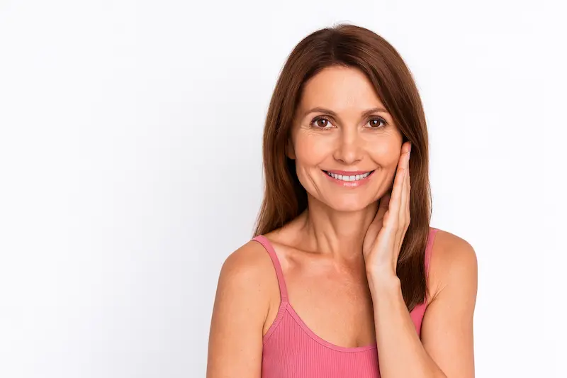 Smiling middle-aged woman with brown hair touching her cheek against a white background, representing natural results after a facelift local anesthesia procedure.