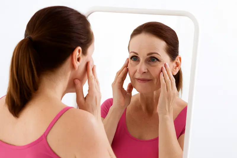 Middle-aged woman examining her face in the mirror, thinking about deep plane facelift in your 40s and early signs of facial ageing.