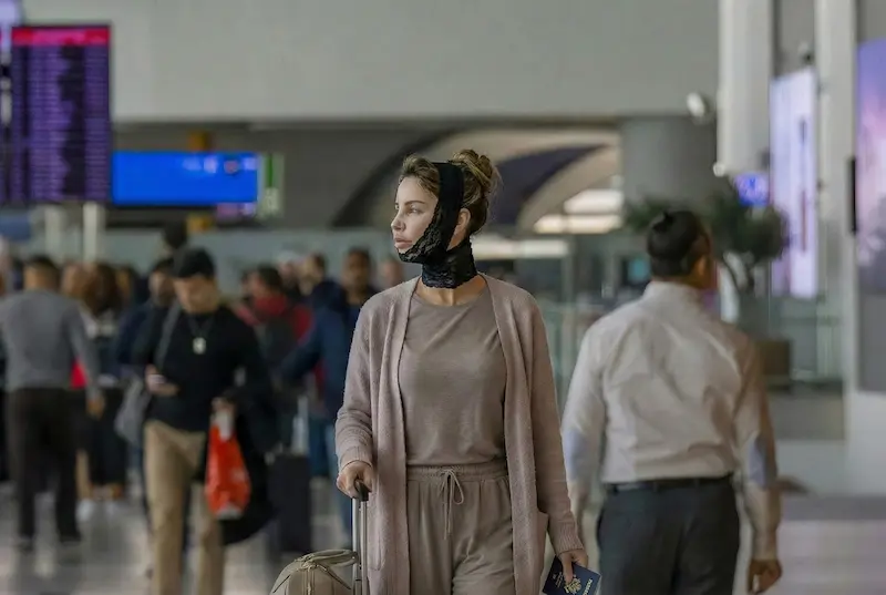How Soon Can You Fly After A Facelift — patient wearing a post-op facelift chin/neck compression wrap while walking through an airport with a suitcase.