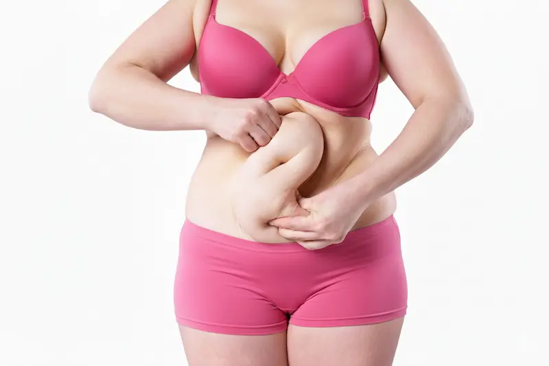 Plus-size woman in pink underwear pinching loose belly skin, symbolizing post-pregnancy body changes and researching Mommy Makeover Cost Turkey.