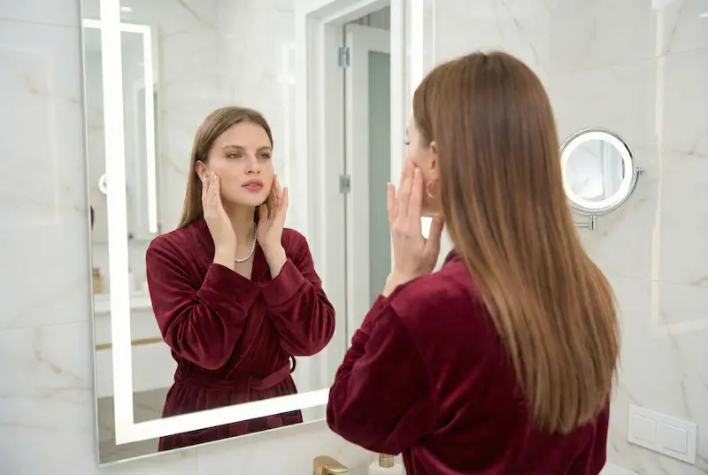 Woman checking her jawline and cheeks in a bathroom mirror, illustrating natural looking facelift turkey goals for subtle facial rejuvenation.