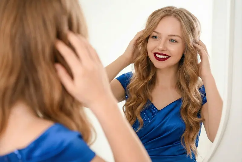 Woman looking in a mirror and touching her hair, representing recovery and results after hair transplant and supporting graft healing.