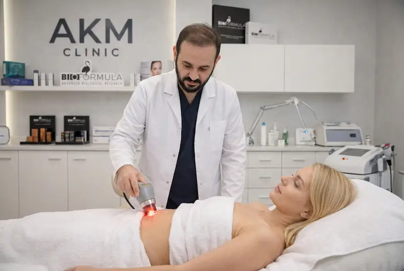 AKM Clinic scar-care protocol session showing Dr. Akif in a white coat using a handheld laser/LLLT device on a patient’s tummy tuck scar in a treatment room.