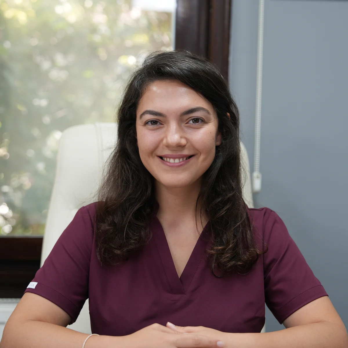 Porträt von Aylin Akbulut Yılmaz in bordeauxroter OP-Kleidung an ihrem Schreibtisch vor einem Fenster