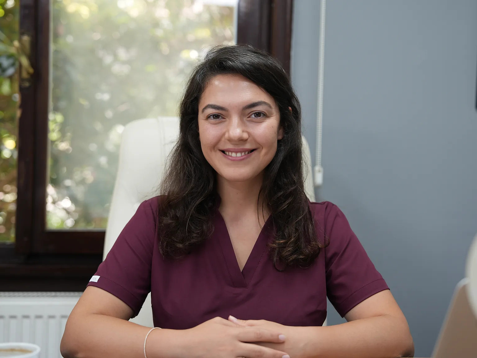 Aylin Akbulut Yılmaz in bordeauxroter OP-Kleidung lächelnd an ihrem Schreibtisch im Sprechzimmer